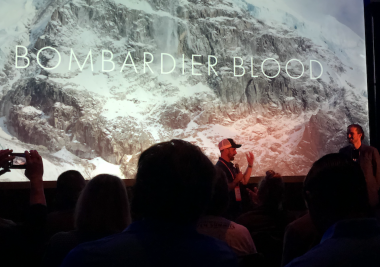 men and hemophilia | Hemophilia News Today | A photo of the premiere of the documentary "Bombardier Blood" at the National Hemophilia Foundation conference in Orlando, Florida. A man is speaking on stage in front of a large screen.