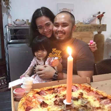 birthday | Hemophilia News Today | Alliah hugs her husband, Jared, while their daughter, Cittie, sits on his lap. They are all smiling for a photo with a pizza and large orange candle on the table in front of them.