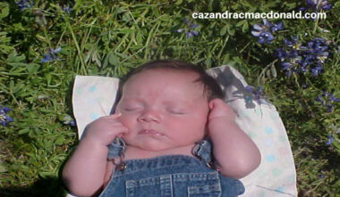 A baby wearing denim overalls lies on his back on a blanket in a field of grass and bluebonnets. His eyes are closed, and he's holding his arms up near his head. 