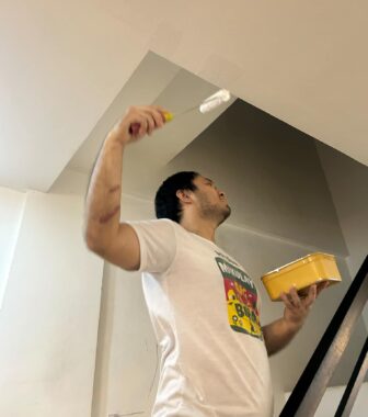 An angular photo taken from below the subject shows a man in a white T-shirt with a paint roller painting a ceiling and some type of ceiling duct. He holds a small, yellow paint tray in his left hand.