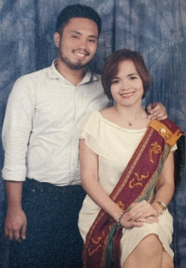 A man and a woman pose before a blue curtain or screen with blue-white designs. He has a short-cropped beard and is standing, wearing a white dress shirt and dark pants. She is seated, seemingly on a stool, in a white dress and a red sash with gold borders and designs. His arm is around her shoulder.