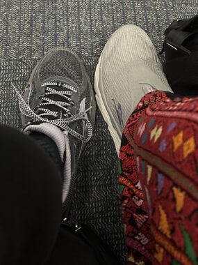 Two people sitting side by side extend a foot for a photo. On the left, we see a person's lower right leg and foot wearing black leggings and a gray and pink sneaker. On the right, a person's left leg and foot sports colorful patterned pants and a white sneaker.