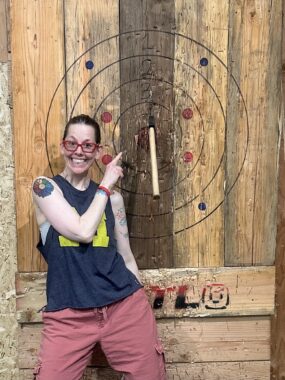 A woman flashes a big smile while standing in front of a wooden panel marked with a bull's-eye. With her right hand, she points over her left shoulder at an ax, embedded almost in the center of the target. She's wearing red-framed glasses and a gray tank top. A colorful tattoo is visible on her right shoulder.