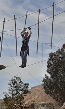 A woman stands on a cable while navigating an aerial ropes course. She's wearing a harness and other safety equipment and is holding on to another cable above her head. 