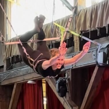 A woman is shown midair, her knees bent around a trapeze bar and her arms outstretched in front of her. She's wearing a harness connected to cables to support her and has a big smile on her face.