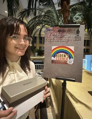 A woman in glasses and wearing a nose ring smiles coyly as she holds a medium-sized white and gold box that holds a fountain pen inside. She's standing next to a small vendor sign that says "Alden's Corner." There are tropical trees behind her.