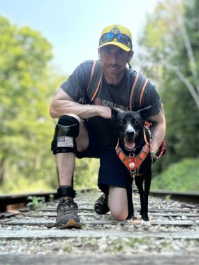 A man kneels down on railroad tracks and poses with his black dog in a forested area.