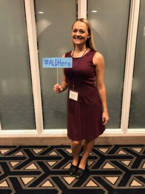 A woman in a wine-colored dress stands in what looks like a hotel hallway or conference center holding a blue sign that says "#ALDHero."