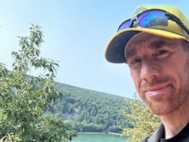 A headshot of a man with a yellow baseball cap and sunglasses while standing in front of a large, forested mountain.