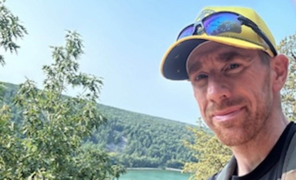 A headshot of a man with a yellow baseball cap and sunglasses while standing in front of a large, forested mountain.