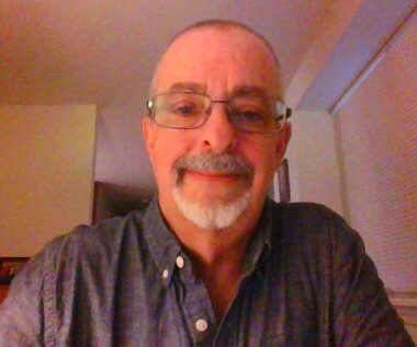 A man in a dark blue, button-up shirt appears in a photo apparently taken from a laptop or computer camera. He's balding, wears glasses, and has a mustache and goatee.
