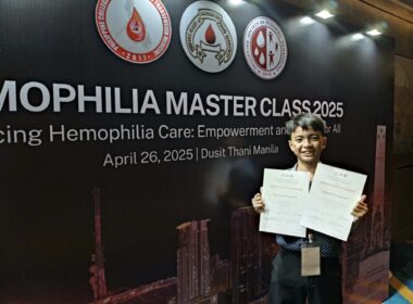 A young man holds up two certificates and smiles proudly while standing in front of a wall that says "Hemophilia Master Class 2025."
