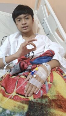 A young man flashes the "OK" sign with his hand as he lies in a hospital bed attached to IVs and covered in a colorful blanket.