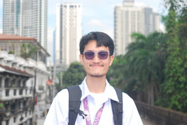 A young, 20-something man stands on a city street with trees and tall buildings in the background. He is wearing nice clothes, sunglasses, and a backpack